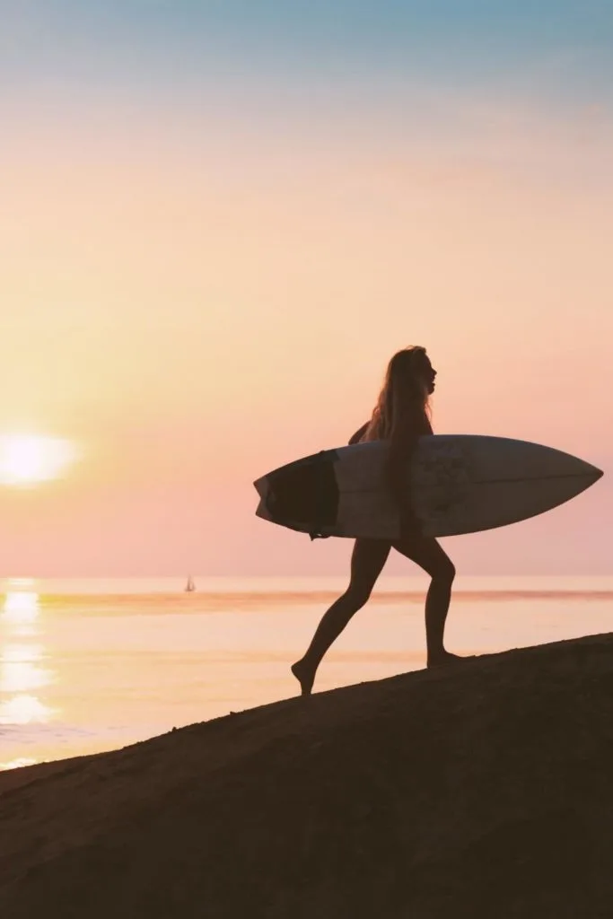 Photo of a woman silhouetted by the sunrise while carrying a surfboard.