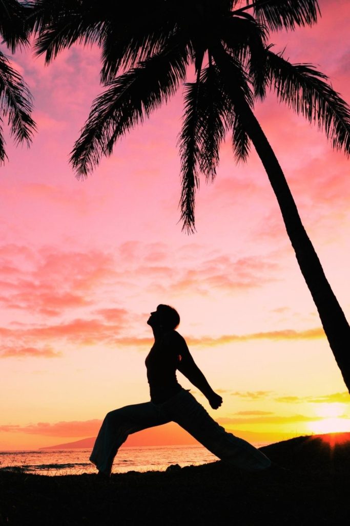 Photo of a person doing yoga during sunset with a palm tree and beach in the background.