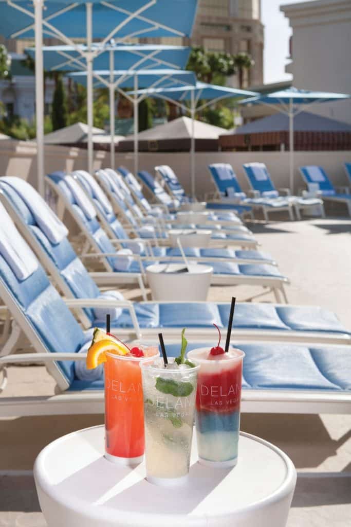 Photo of a row of pool lounge chairs with a table full of colorful cocktails that say "Delano Beach Club" on the cups.