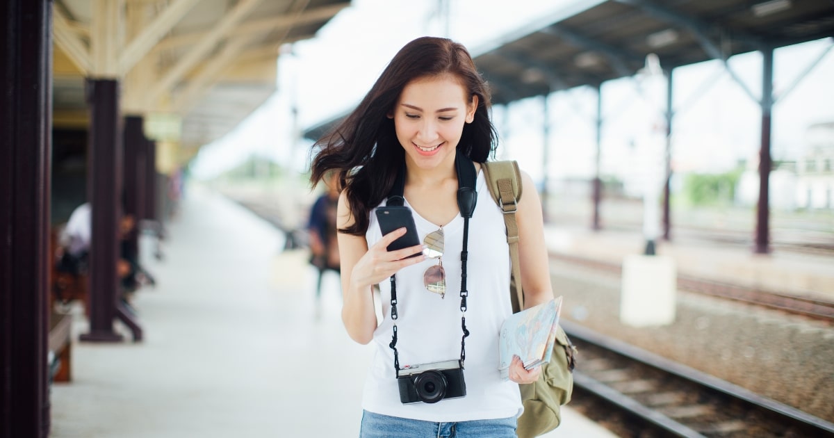 Photo of a young woman at a train station looking at her phone while carrying a camera and a map.