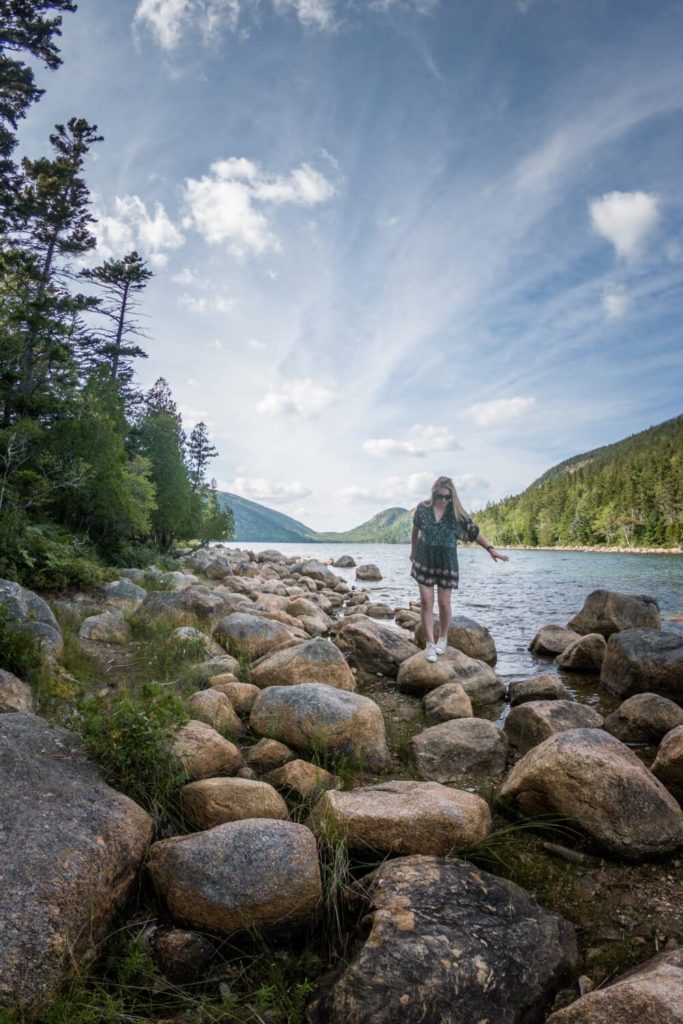 Photo of a woman walking across large rocks along the shoreline of Jordan Pond.