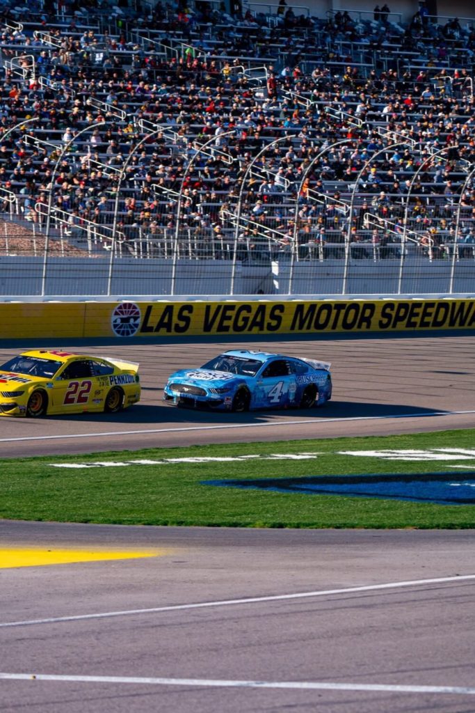 Photo of a NASCAR race at Las Vegas Motor Speedway with a blue and red car racing by in front of a crowd in the background.