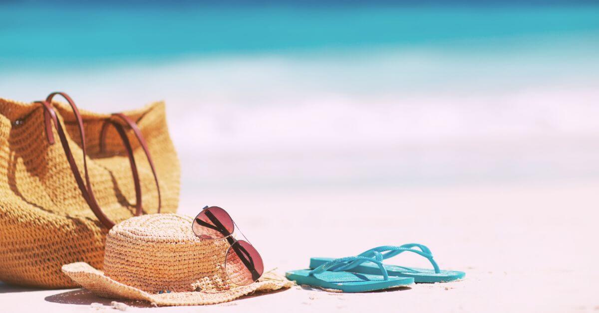 Photo of a straw beach bag, straw hat, sunglasses, and flip flops sitting on a sandy beach.