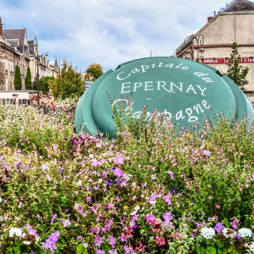 Closeup photo of a giant champagne cap sitting amongst flowers that says: Epernay: Capitale du Champagne.