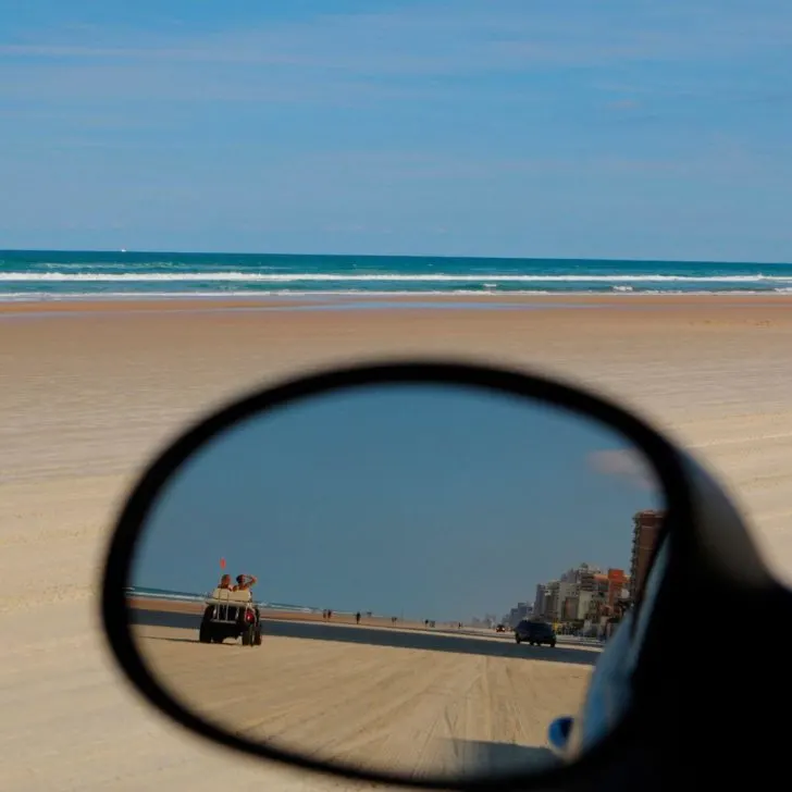 Photo of a reflection in a car's side-view mirror of people driving on the hard sand in Daytona Beach, Florida.