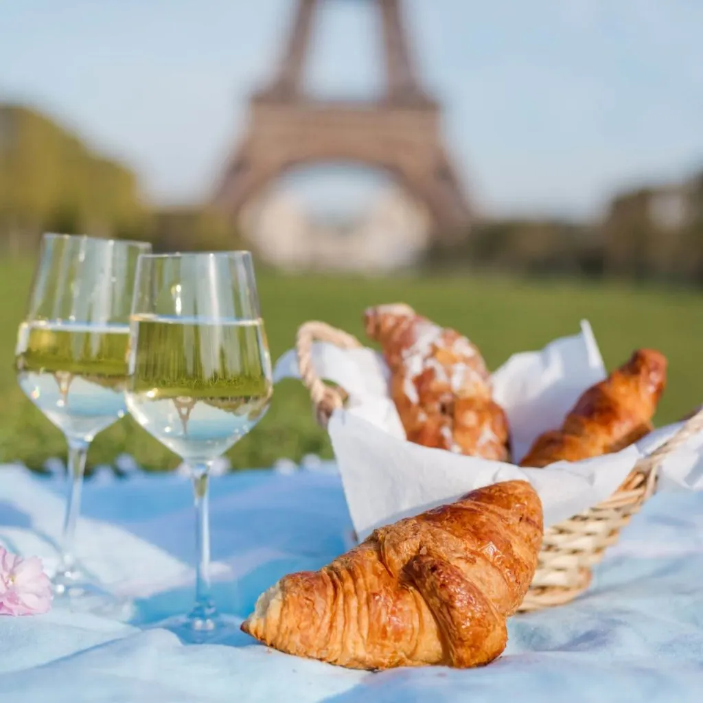 Closeup of a basket of croissants and 2 wine glasses on a picnic blanket with the Eiffel Tower in the background.