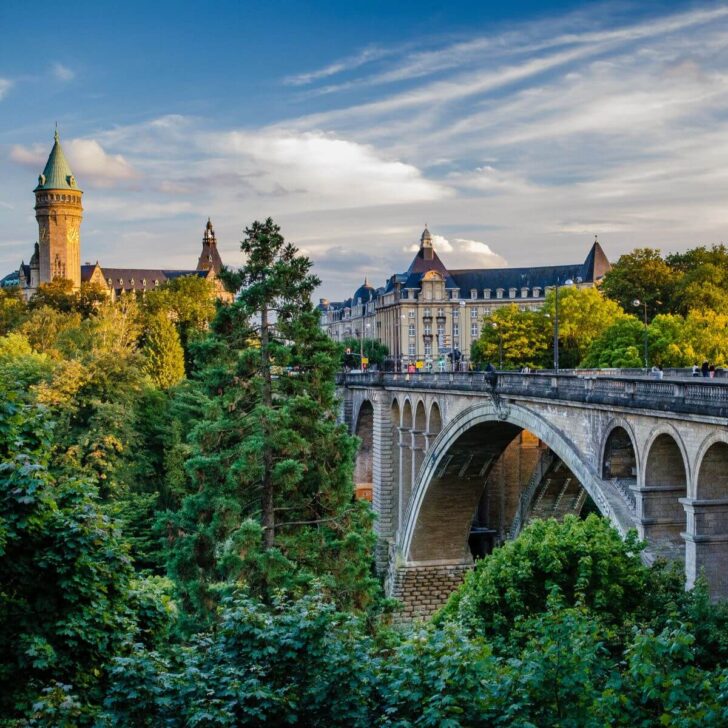 Photo of a beautiful arch bridge and a castle-like building with trees in the foreground in Luxembourg City.