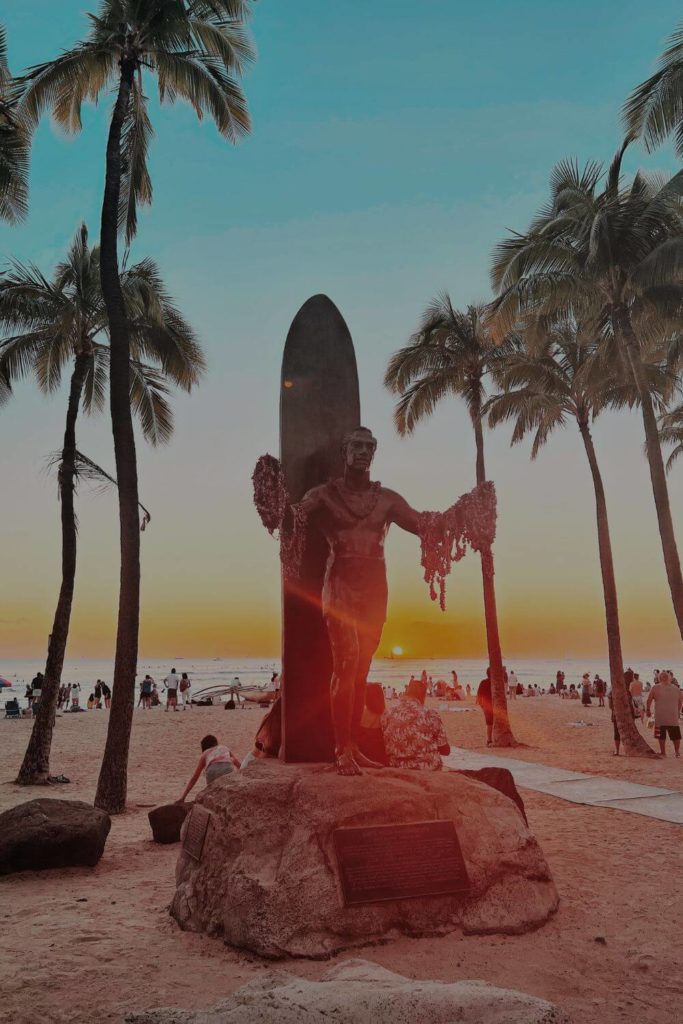 Photo of the Duke Kahanamoku statue in Waikiki Beach in Oahu, Hawaii while the sun sets in the background.