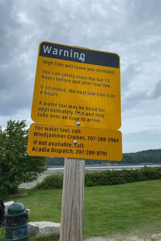 Photo of a yellow warning sign at the start of the Bar Island Trail with information on what to do if you get stranded on the island due to high tide.
