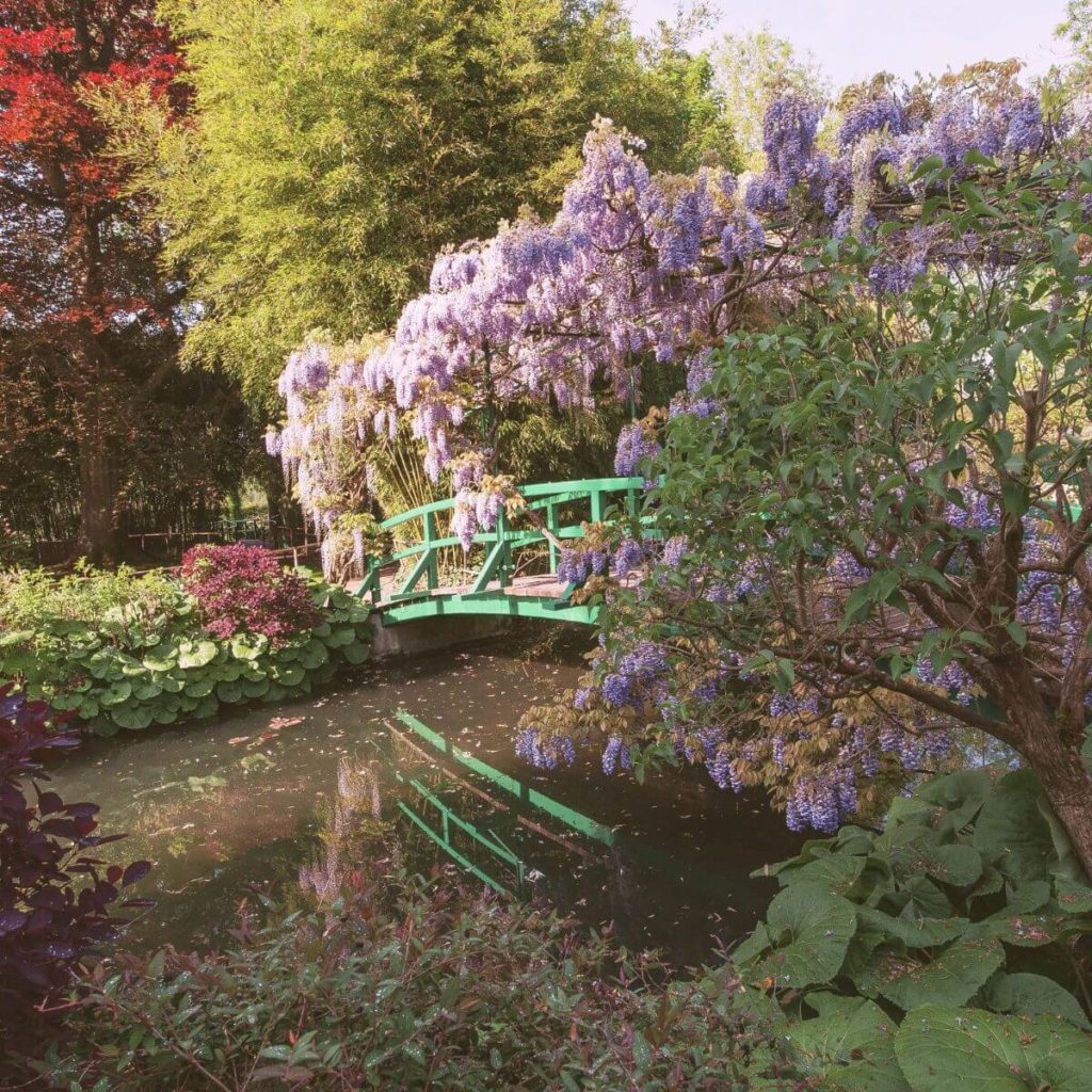Photo of part of the gardens at Monet's former home in Giverny, France, with a quaint green bridge over a pond surrounded by purple flowers.