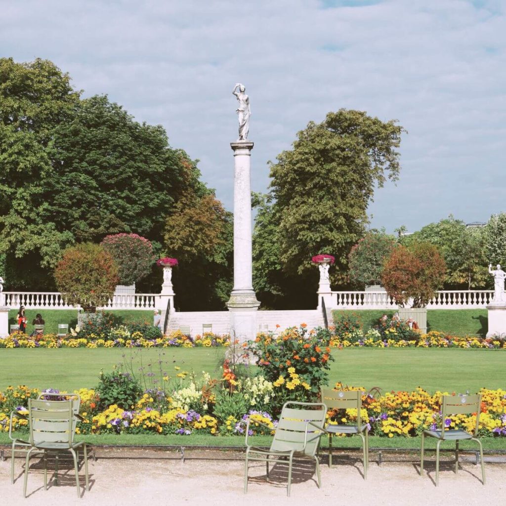 Photo of Luxembourg Gardens with chairs along a sandy path in the foreground and lots of bright flowers and lush green trees throughout the background.