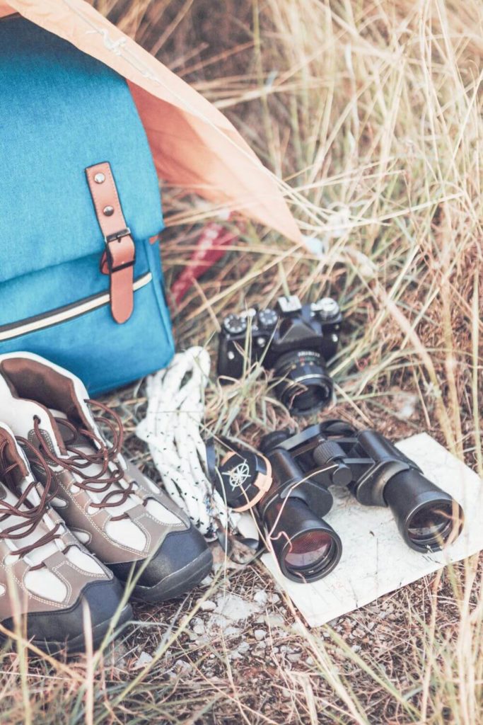 Photo of a backpack, trail boots, camera, binoculars and other outdoor gear on top of some grass.