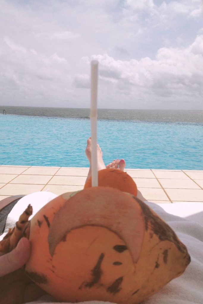 Photo of a coconut with a straw in it on a woman's lap while she lounges next to a pool overlooking the Indian Ocean in Bentota, Sri Lanka.