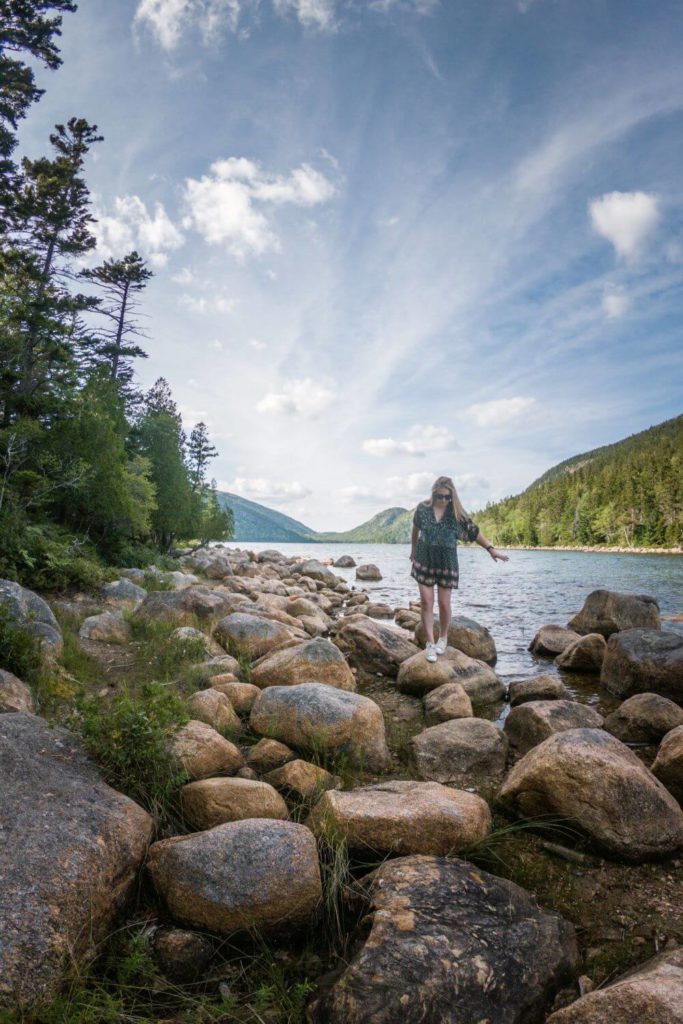 Photo of a woman walking along large rocks that line the shore of Jordan Pond.