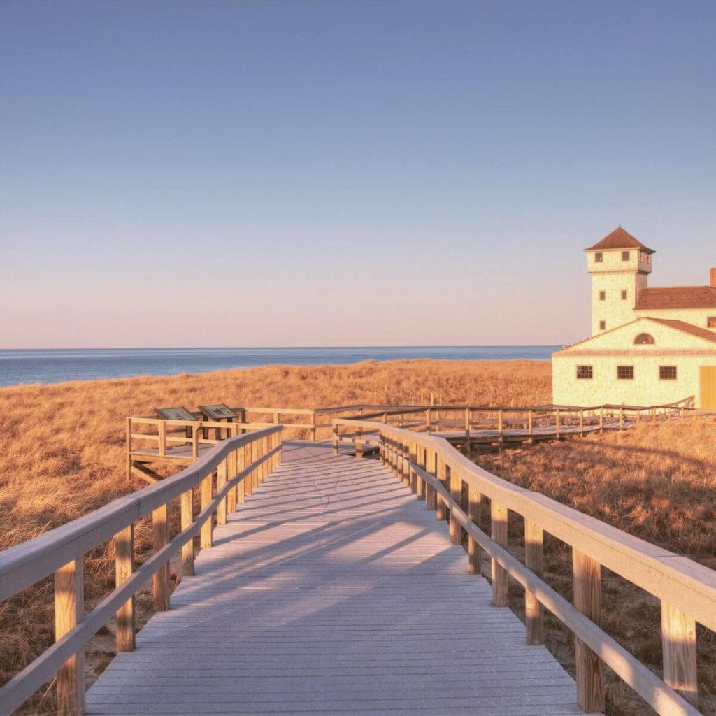 Photo of a wooden walkway leading to an old lifeguard station surrounded by sea grass with Race Point Beach in Provincetown, MA, in the background.