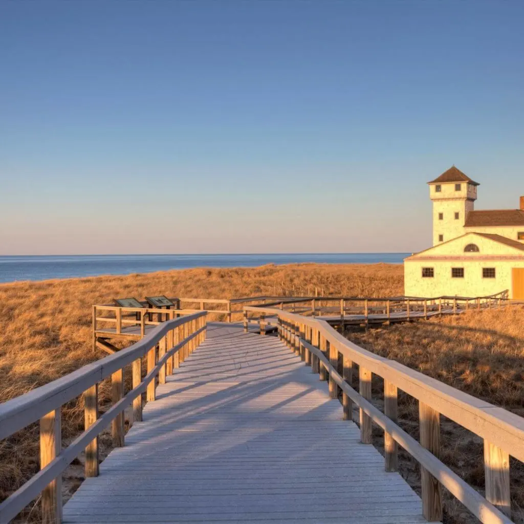 Photo of a wooden walkway leading to an old lifeguard station surrounded by sea grass with Race Point Beach in Provincetown, MA, in the background.