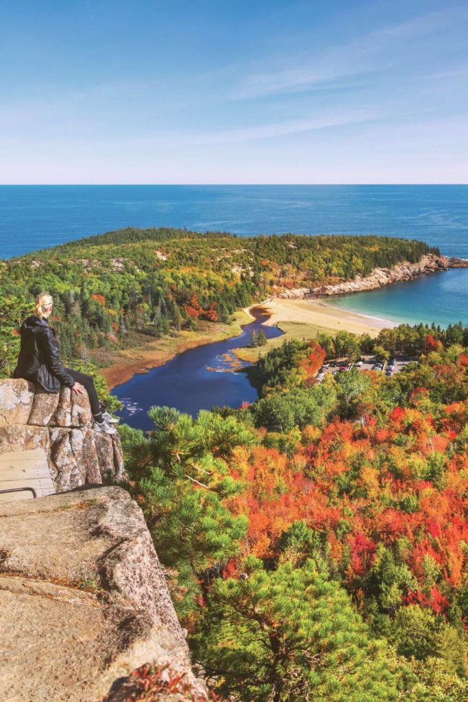 Photo of a woman sitting on the ledge of a rock looking out over Sand Beach and the forest below her from the summit of the Beehive hike.