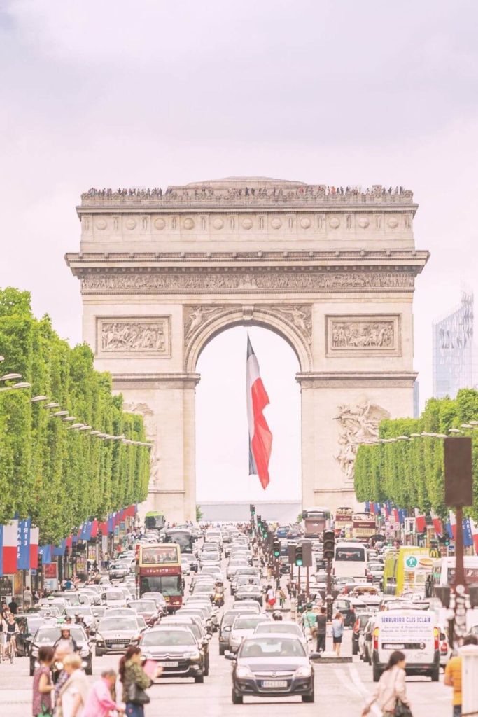 Photo of the Arc de Triomphe with the French flag hanging from the middle of the arch and traffic in the foreground.