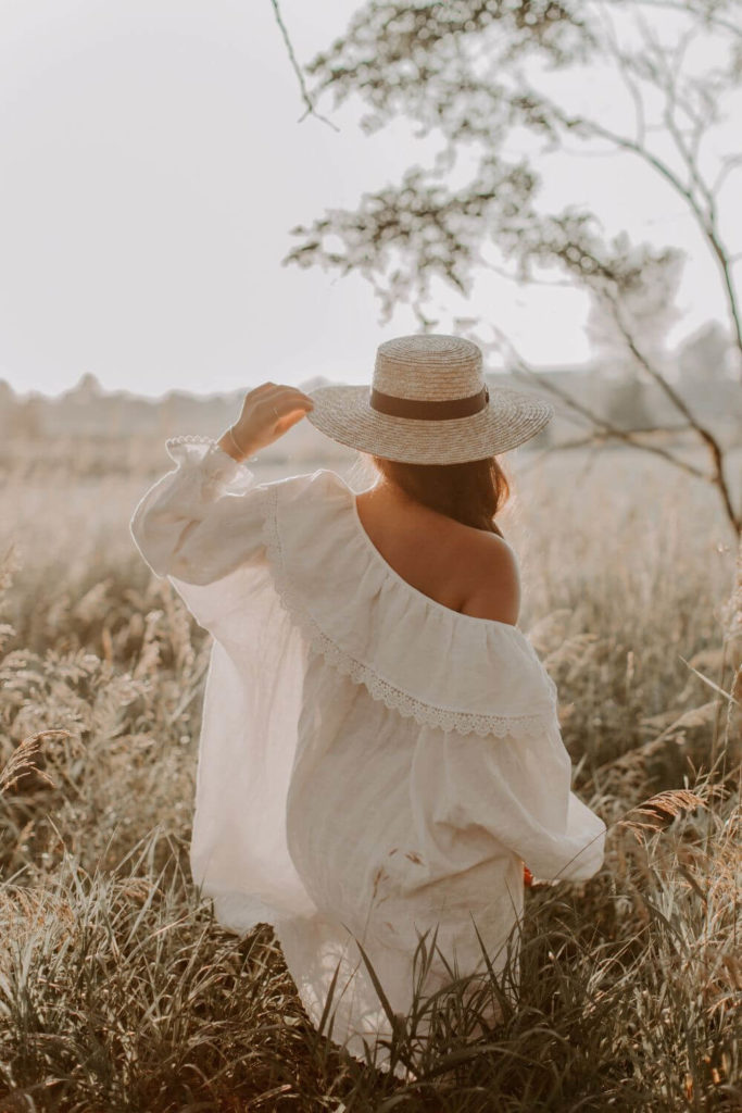 Photo of a woman wearing an oversized white ruffled boho dress and and straw hat while frolicking in a field.