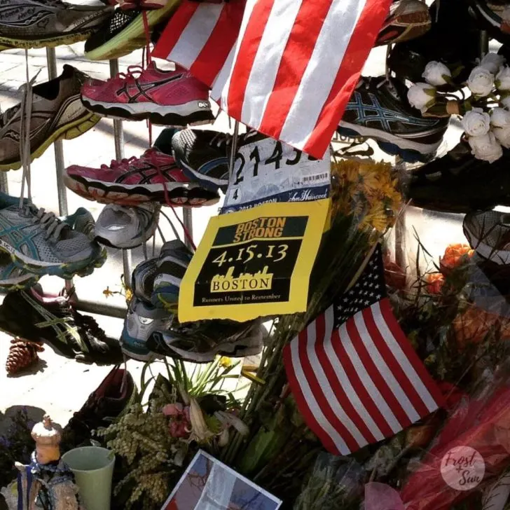 Closeup photo of a makeshift memorial for the 2013 Boston Marathon bombing with runners' sneakers, race bibs, flowers, and American flags tied to security barriers.