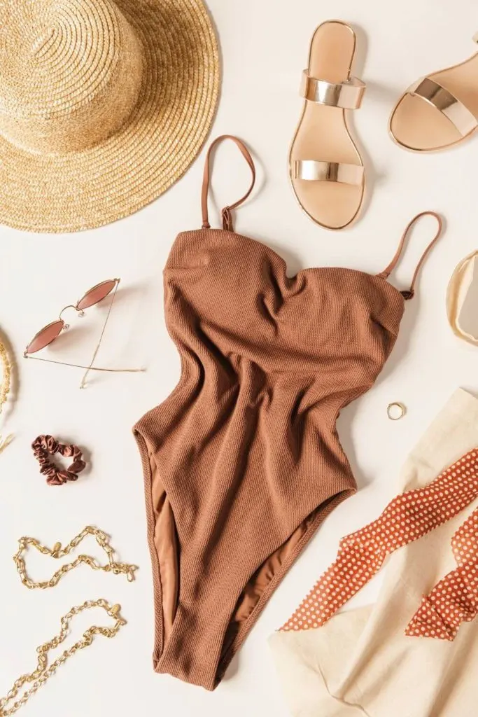Flat lay photo with neutral color theme: gold sandals, straw hat, gold jewelry, cocoa colored swimsuit, and more.