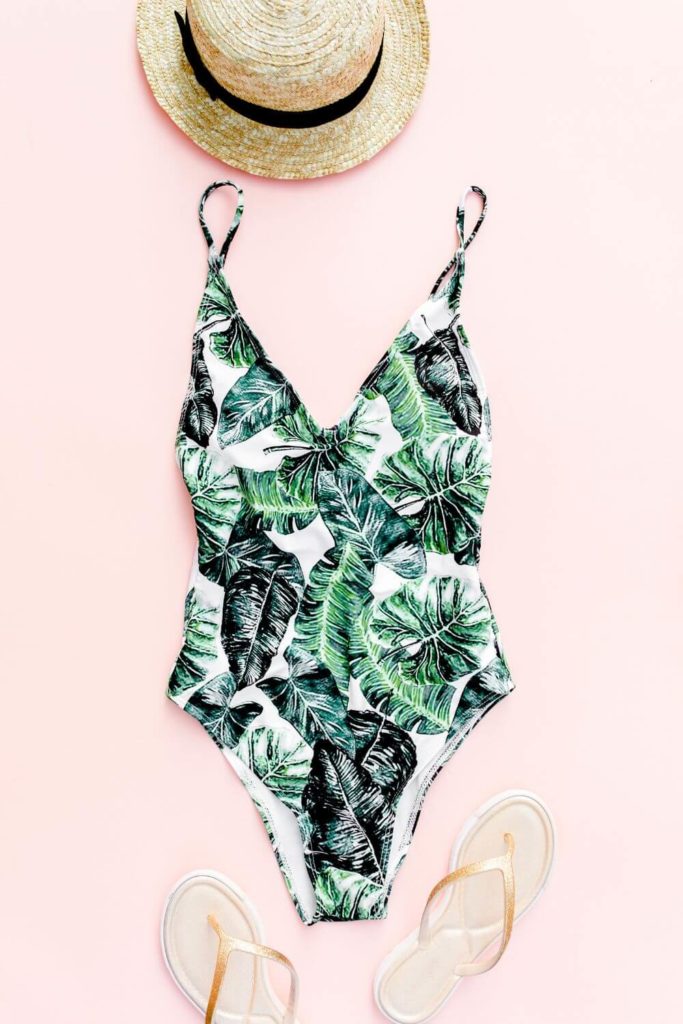Flat flay photo of a straw hat, tropical print one piece swimsuit, and gold flip flops.