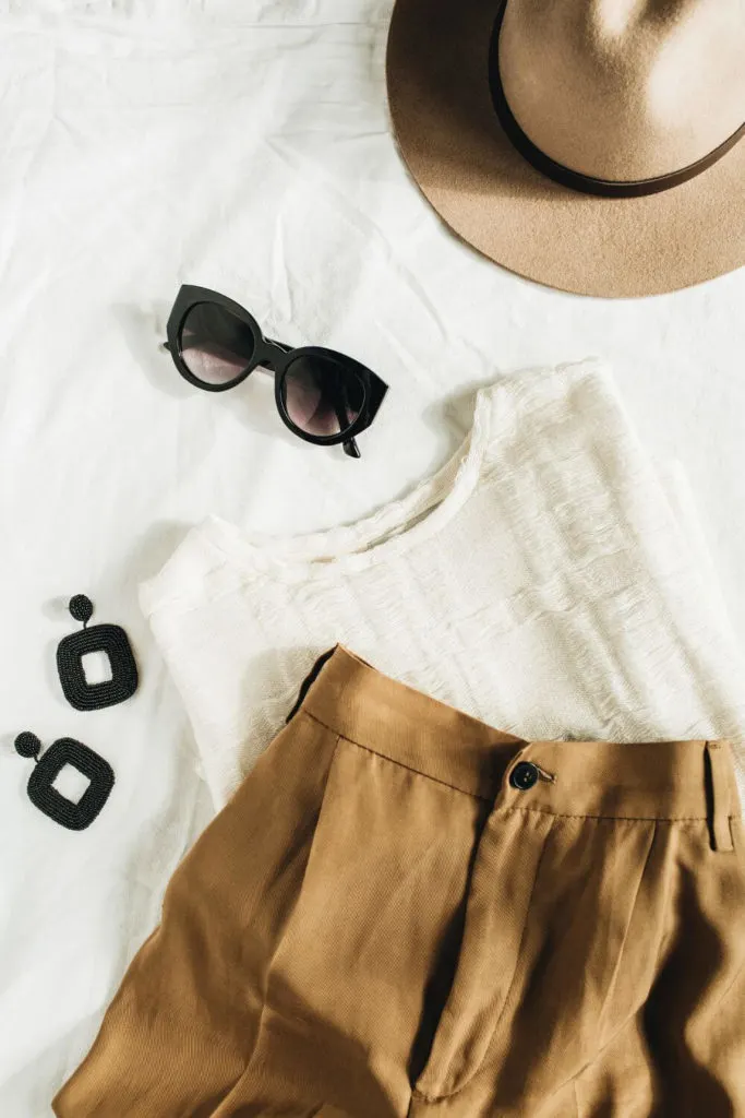 Flat lay photo with a white linen shirt and khaki dress shorts, black earrings, black cat eye sunglasses, and a tan hat.