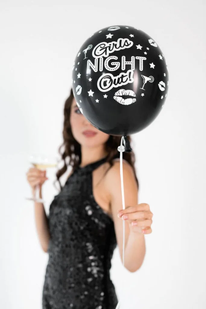 Photo of a woman in a black sequin dress with a glass of champagne in one hand and a balloon that says "Girls Night Out" in the other.