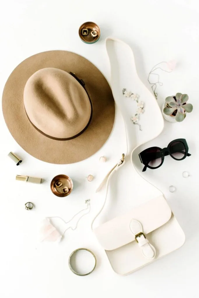Flat lay photo with a white shoulder bag, black cateye sunglasses, jewelry, makeup, and a tan hat.