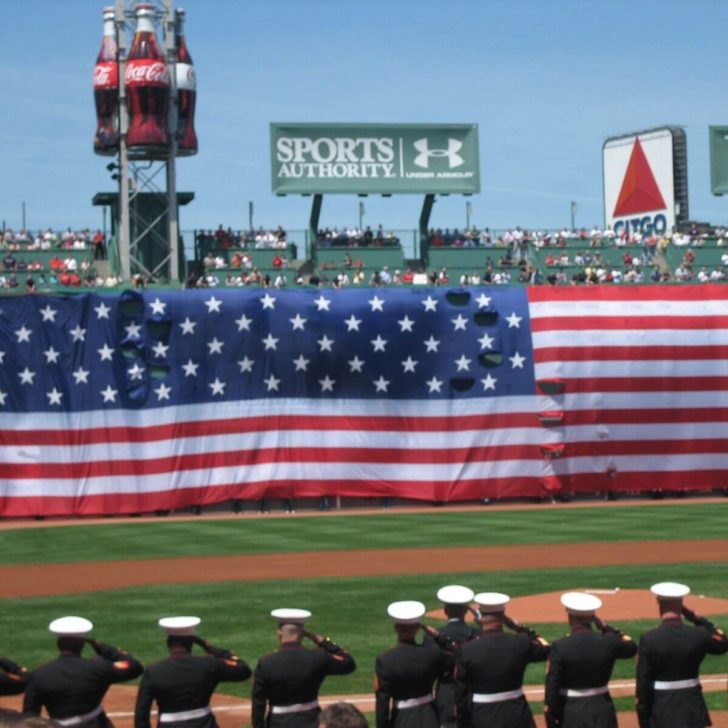 Photo of a flag draped across the Green Monster at Boston's Fenway Park with US Marines in dress uniform saluting in the foreground.