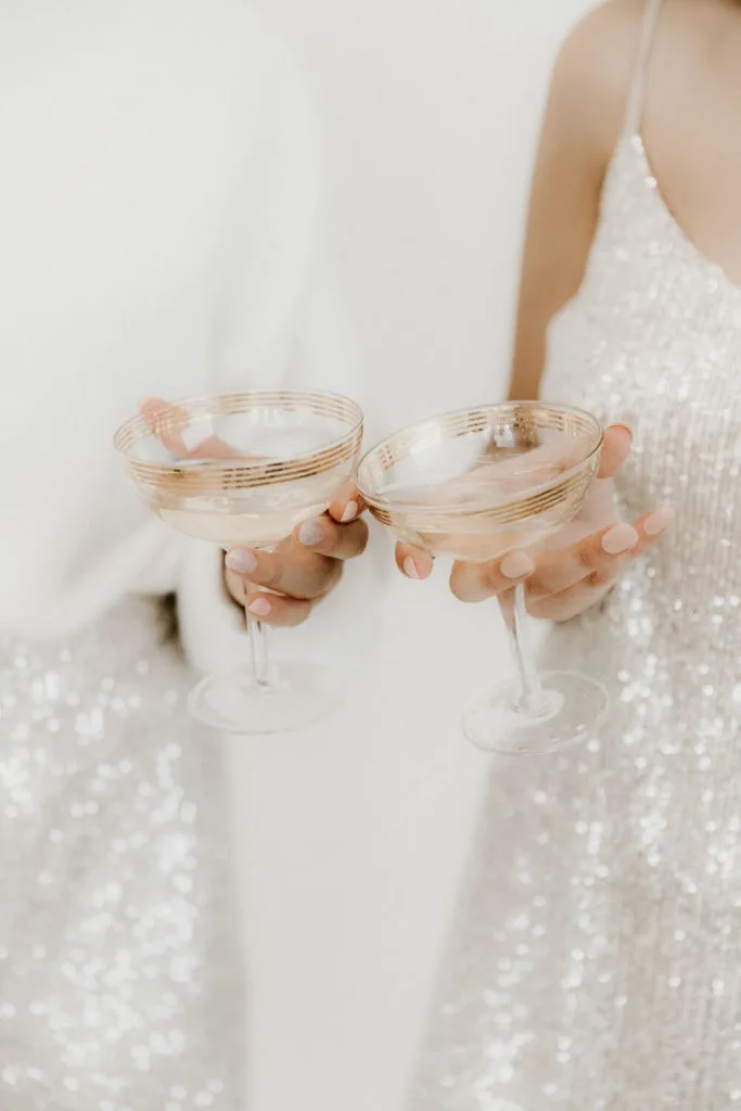 Closeup of 2 women wearing white and silver sequin dresses, clinking 2 glasses of champagne.
