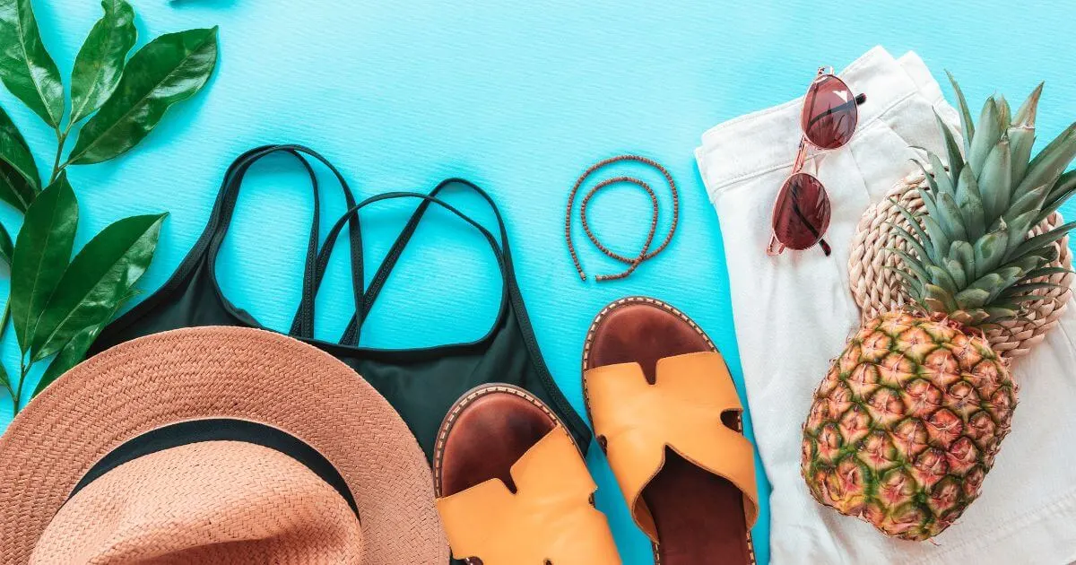 Flat lay photo of a pineapple, straw hat, flat sandals, sunglasses, black swimsuit, and white shorts.