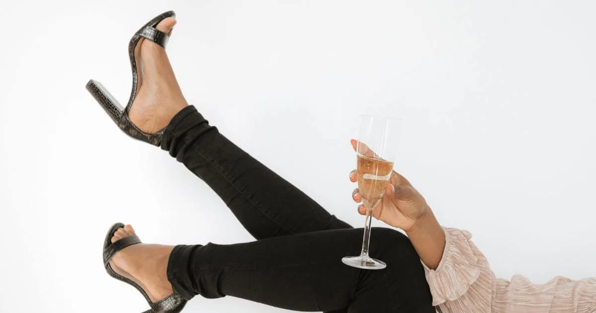 Closeup photo of a woman with her feet in the air, wearing black jeans and black snakeskin heels, holding a glass of champagne.