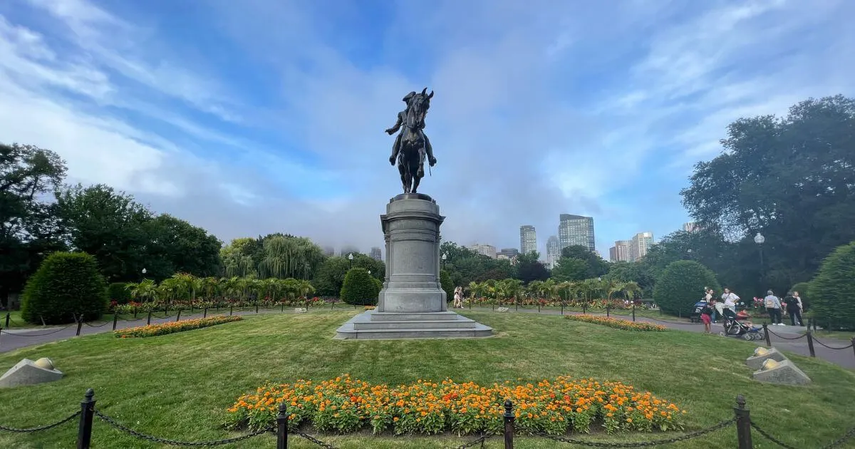 Horizontal photo of the Paul Revere statue in the Boston Public Garden with orange flowers in the foreground and fog rolling into the city in the background.
