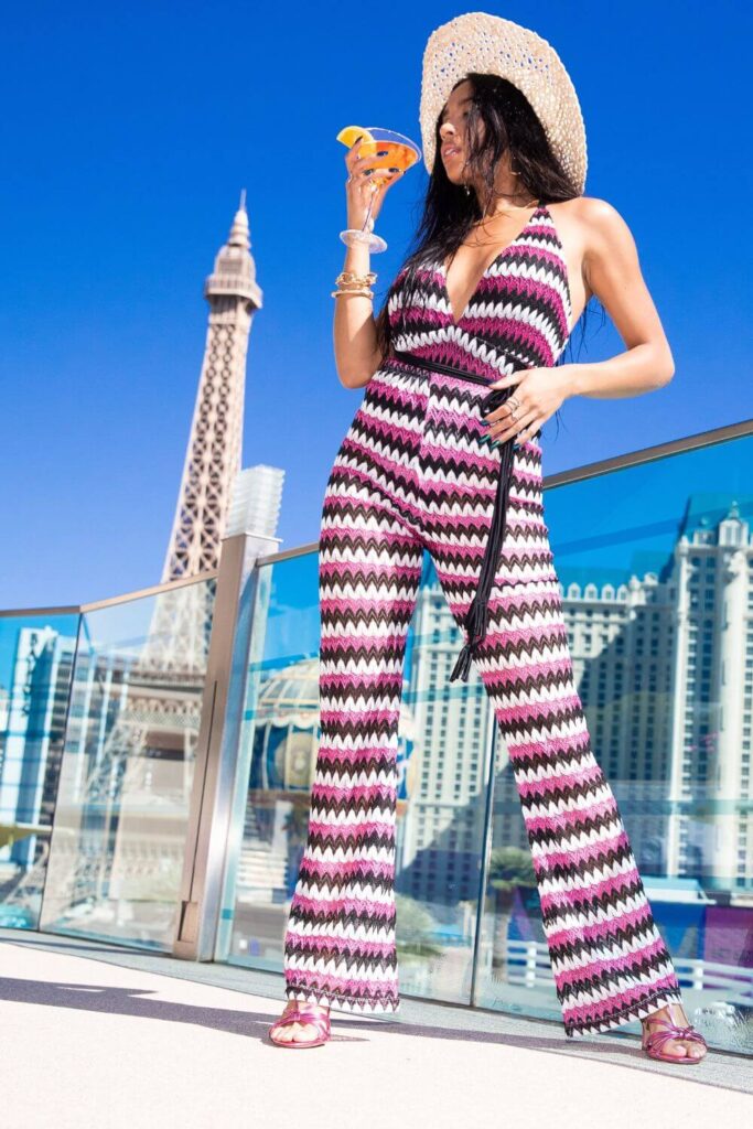 Photo of a woman wearing a striped jumpsuit and sipping a cocktail with the Vegas skyline in the background.