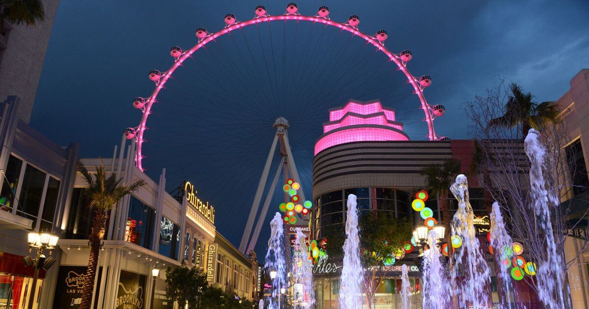 Photo of the LINQ Promenade at night with the fountain in the foreground and the High Roller lit up in pink in the background.