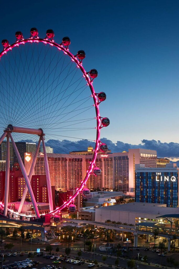 Photo of the High Roller at night, lit up in pink, with the strip behind it.