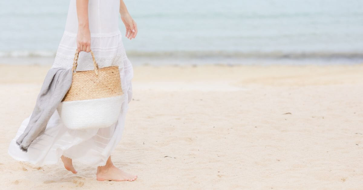 Photo of a woman in a white maxi dress and carrying a straw tote while walking down the beach barefoot.