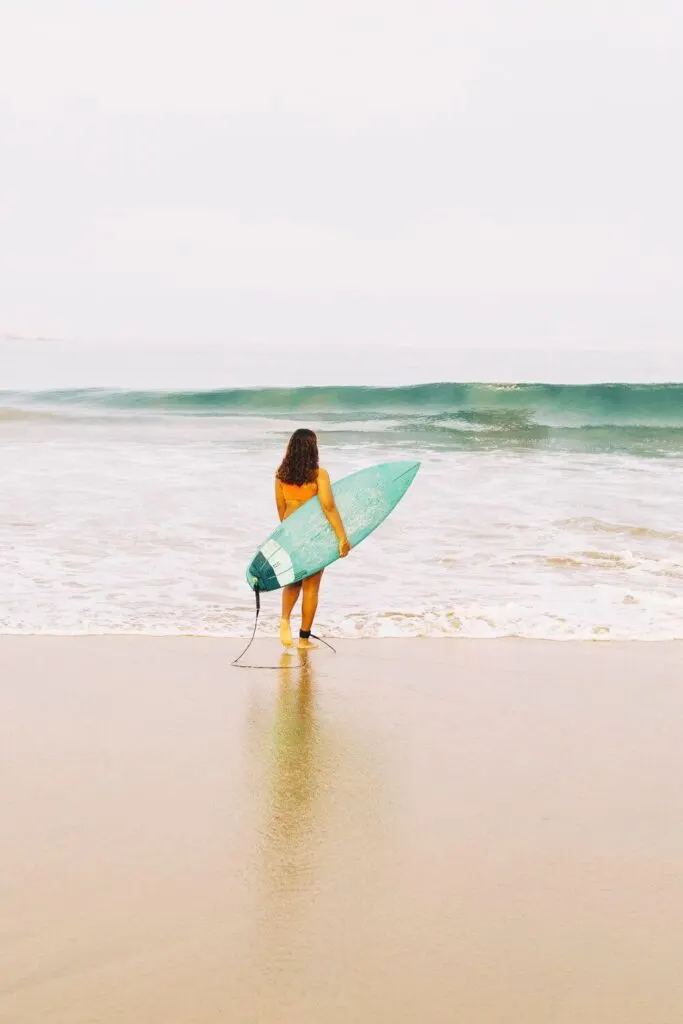Photo of a woman walking toward the ocean while carrying a turquoise surfboard.