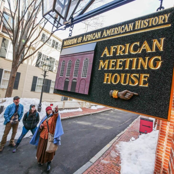 Photo of a tour guide dressed in costume pointing to a sign for the Museum of African American History in Boston, as tour guests look on.