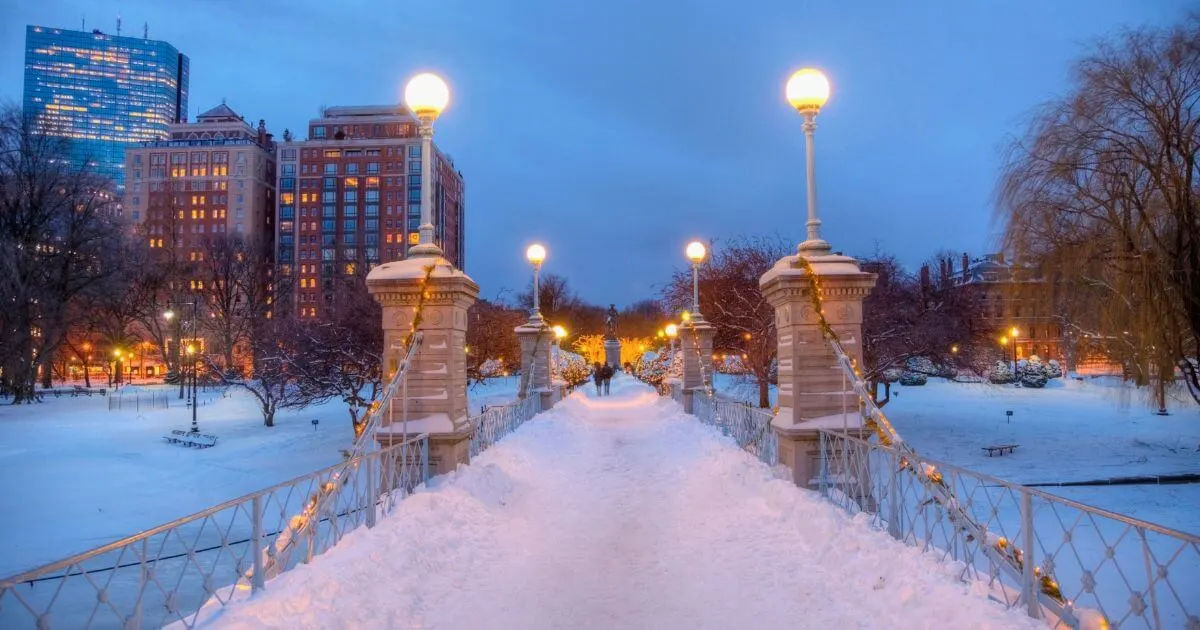 Photo of the foot bridge in the Boston Public Garden at night, with lights lit up and fresh snow on the ground.